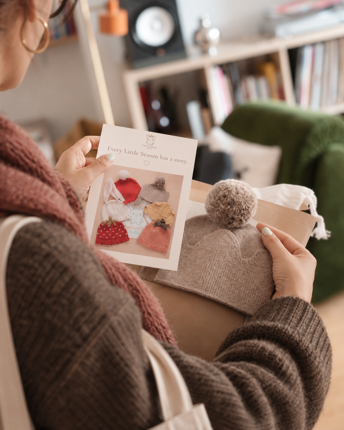 Une personne tient une carte de collection Tiny & Wild et un bonnet Crown tricoté en ouvrant un colis en papier kraft chez elle