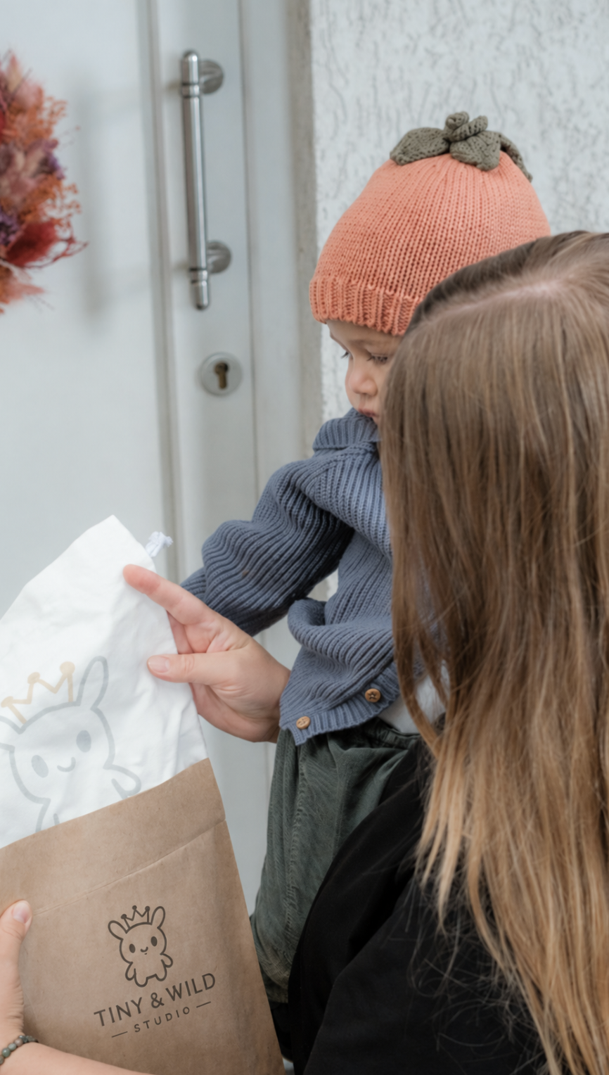Un parent tient un bébé portant le bonnet Pumpkin à côté d’un pochon cadeau Tiny & Wild devant la porte