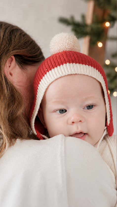 Baby wearing a red and cream Christmas Beanie while resting on a parent’s shoulder beside softly lit greenery