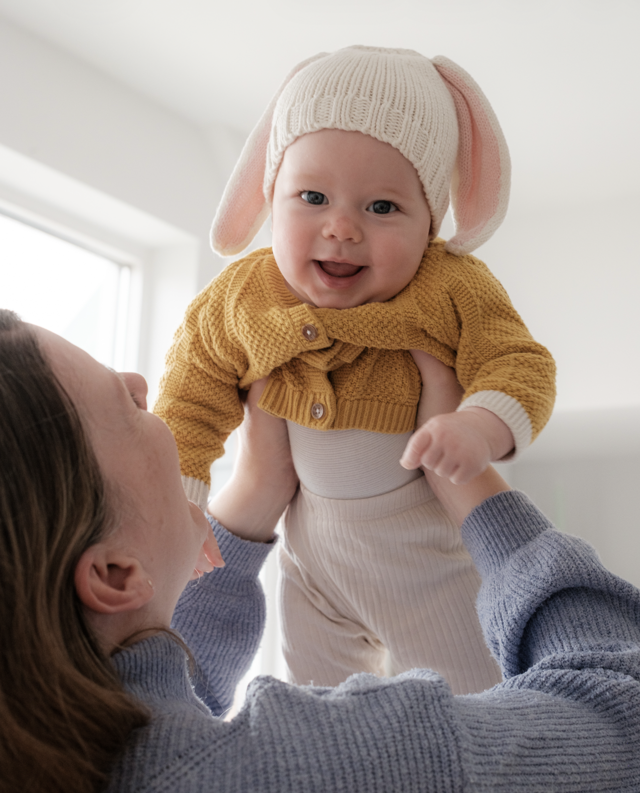 Parent lifting a smiling baby wearing a cream Bunny beanie with pink-lined ears and a mustard cardigan