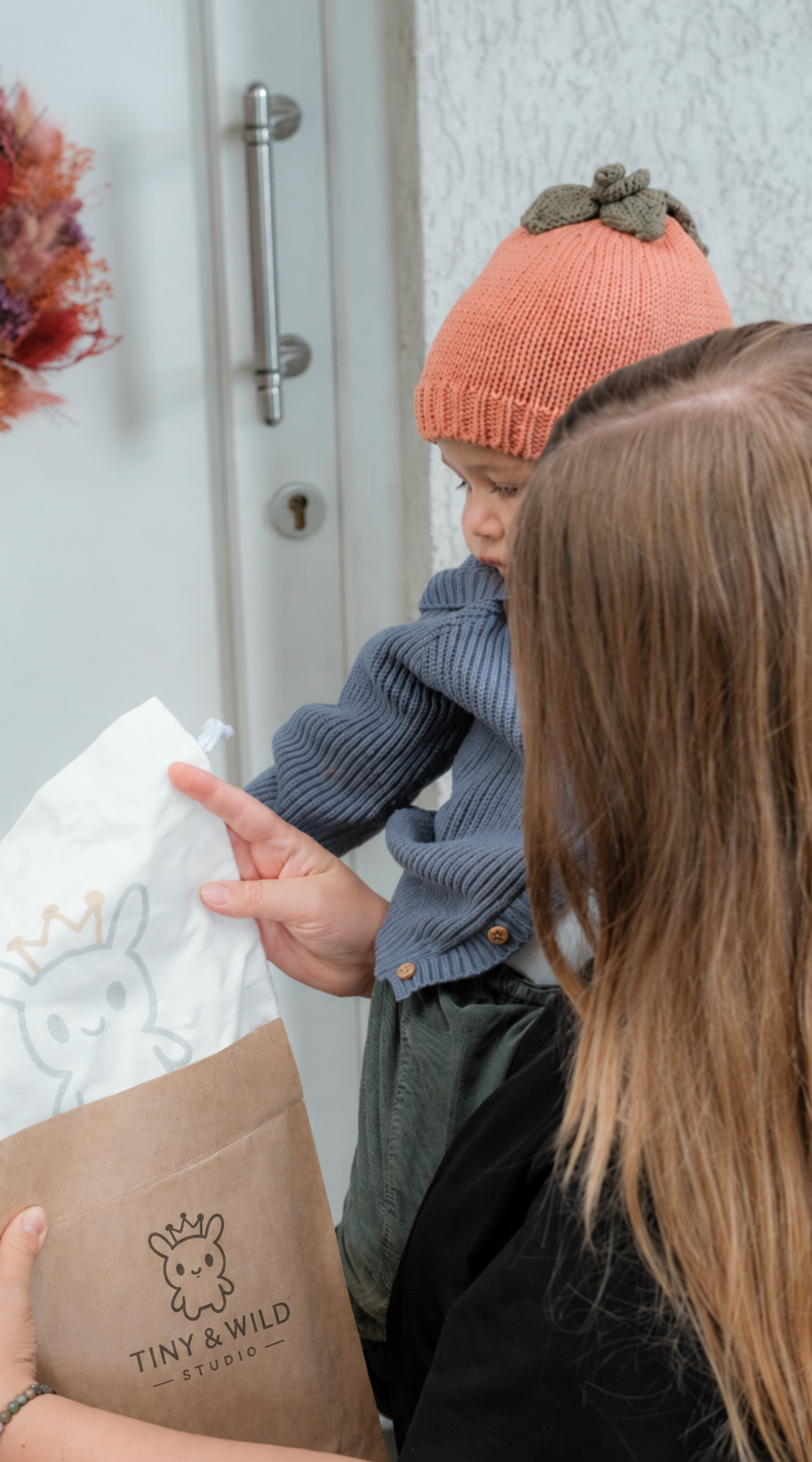 Parent and baby wearing the Pumpkin beanie opening a Tiny & Wild package at the front door