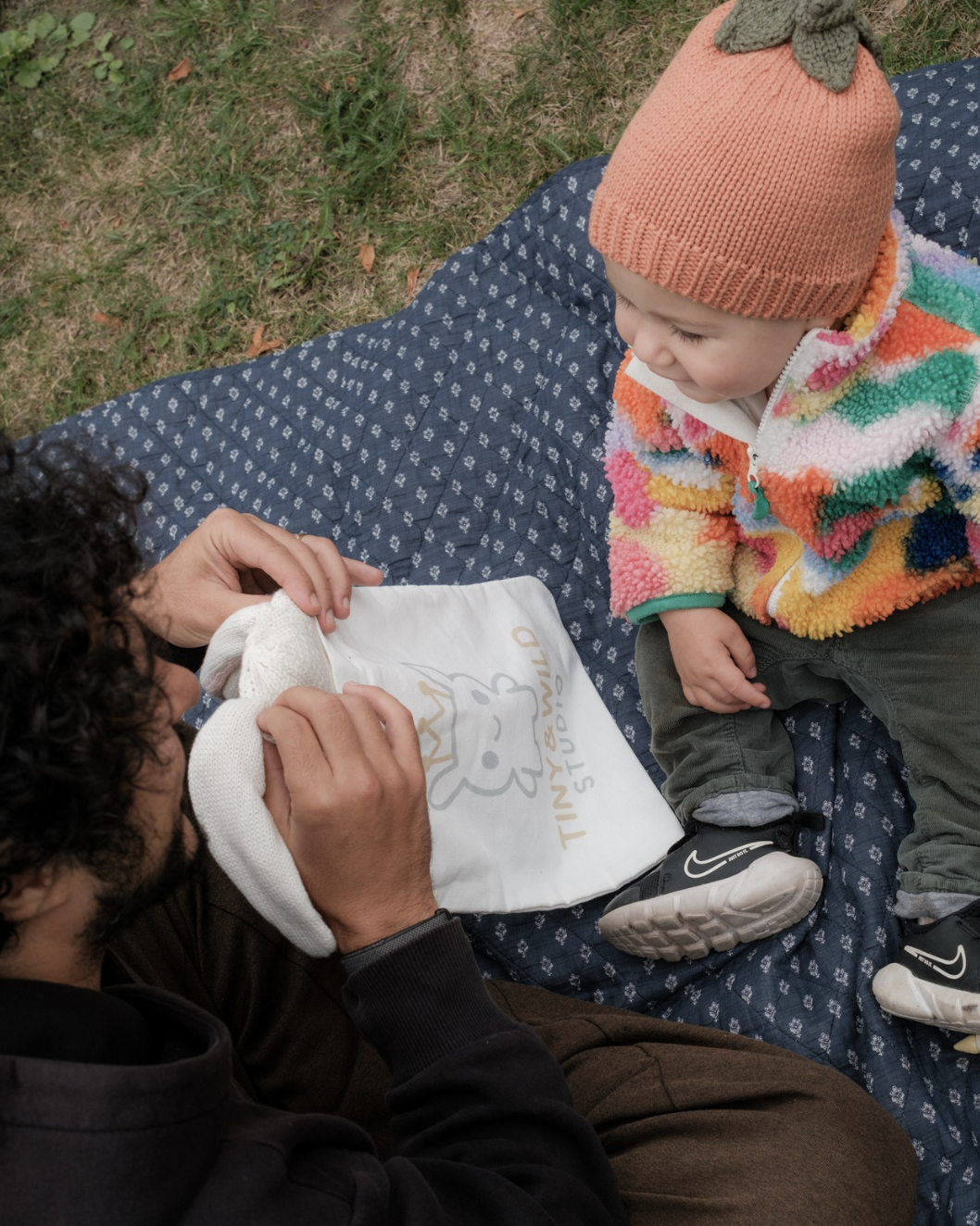 Parent unpacking the Bunny beanie beside a baby wearing the Pumpkin beanie and a Tiny & Wild fabric bag