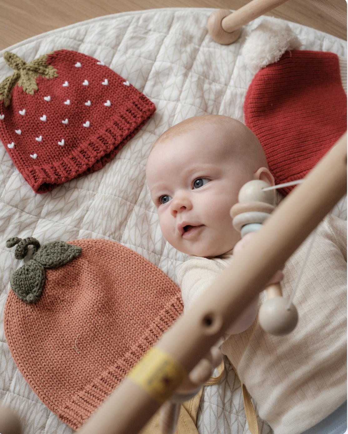 Baby lying beside the Strawberry, Pumpkin and Christmas beanies under a wooden play gym